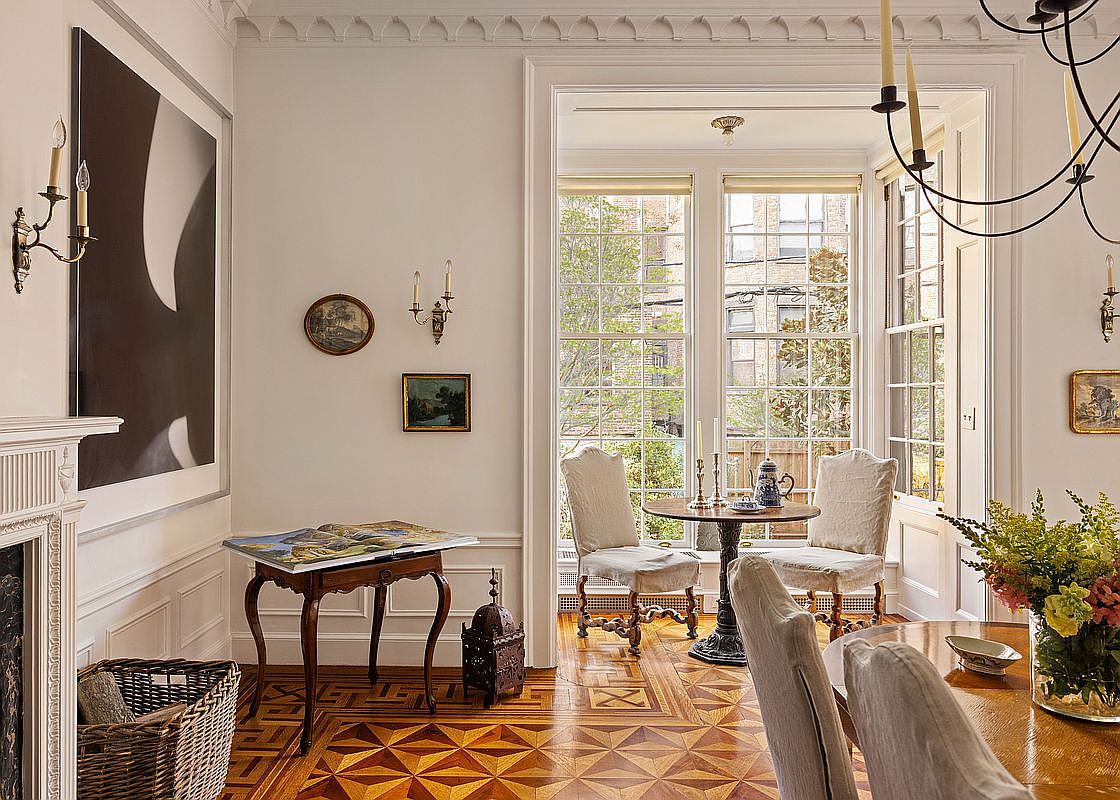 This elegant dining room features intricate parquet wood flooring and classic architectural molding, creating a sophisticated and timeless atmosphere. The space is anchored by a large dining table and a cozy breakfast nook by the window, complemented by tasteful wall art and warm lighting fixtures. The perspective is a wide, eye-level shot that captures the seamless blend of formal dining and casual seating areas.