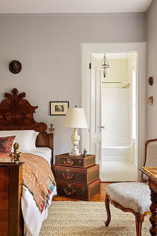 This charming bedroom features a classic, antique-inspired aesthetic with a prominent wooden headboard and a stack of vintage-style trunks serving as a unique nightstand. The room is accented by a patterned throw blanket, a traditional table lamp, and a view into an adjacent white-tiled bathroom, creating a cozy and curated atmosphere. The perspective is a medium shot from the side of the bed, highlighting the blend of vintage furniture and neutral wall tones.