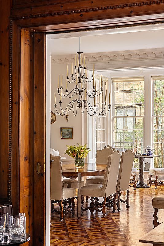 This elegant dining room is framed by rich, dark wood trim, creating a sophisticated transition from the foreground. The space features a classic round wooden table surrounded by upholstered chairs, illuminated by a grand, multi-tiered candle-style chandelier. Large windows allow natural light to flood the room, highlighting the intricate geometric parquet flooring and providing a view of the lush greenery outside.