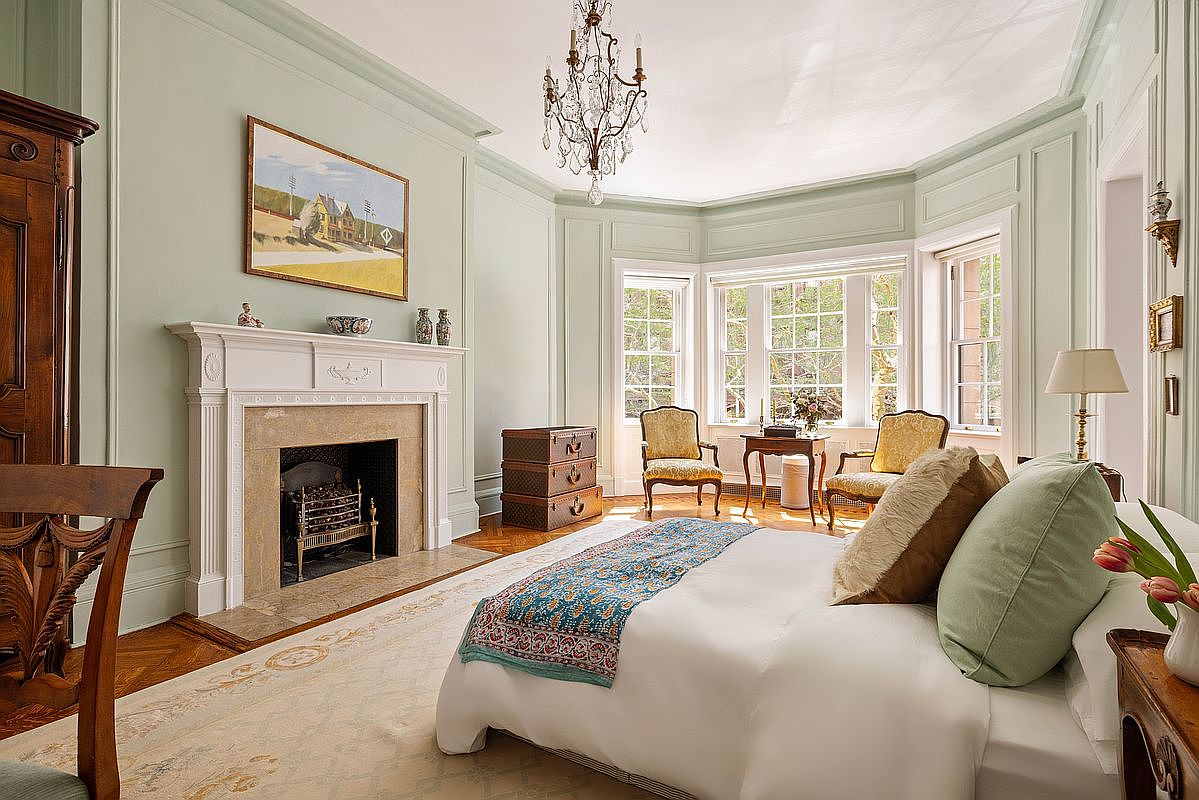 This elegant primary bedroom features a sophisticated light sage color palette, classic crown molding, and a grand fireplace with a marble hearth. The room is bathed in natural light from a large bay window, which frames a cozy seating area with two upholstered chairs, while a crystal chandelier adds a touch of luxury above the plush bed. The perspective is from the foot of the bed, capturing the harmonious blend of traditional architectural details and refined, comfortable furnishings.
