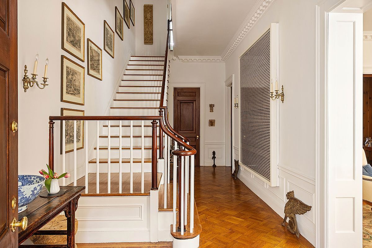 This elegant foyer features a grand staircase with a rich wood handrail and white balusters, set against a backdrop of classic architectural details. The space is adorned with a gallery wall of framed artwork, a polished herringbone hardwood floor, and a sophisticated console table with a decorative bowl. The perspective captures the inviting transition from the entryway into the home's interior, highlighting a refined and traditional aesthetic.