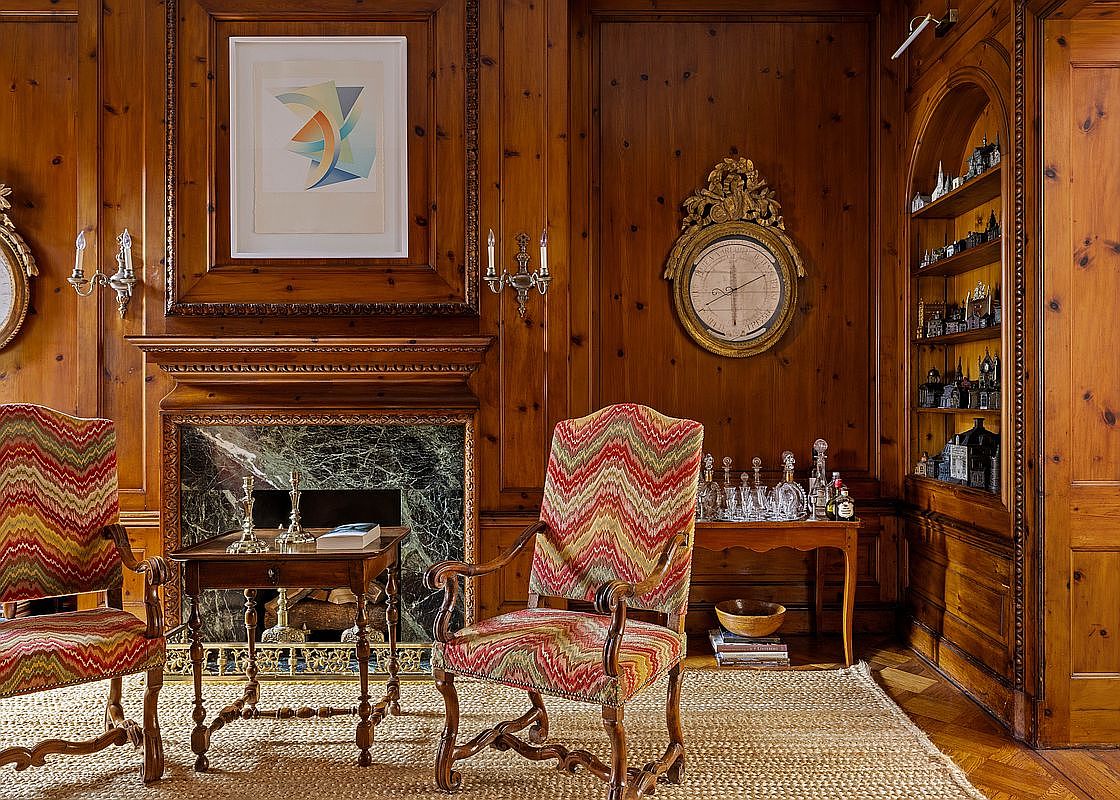 This sophisticated living room features rich, wood-paneled walls and a classic fireplace with a dark green marble surround. Two patterned, chevron-style armchairs are positioned around a small wooden side table, while a built-in arched bookshelf displays a collection of miniature buildings. The room exudes a traditional, scholarly atmosphere, enhanced by elegant wall sconces and a decorative circular barometer.