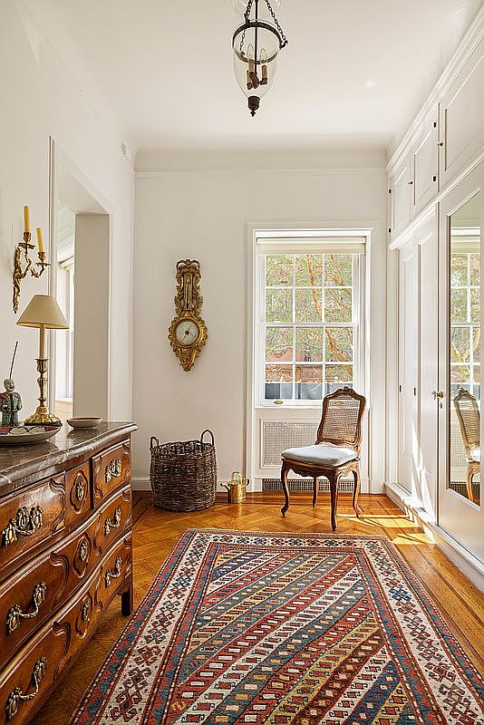 This elegant hallway features warm hardwood flooring covered by a vibrant, patterned area rug and is illuminated by a classic hanging lantern fixture. To the left, an ornate antique wooden dresser topped with a lamp and wall sconces adds a touch of traditional charm, while a window at the end of the hall provides natural light and a view of the outdoors. The space is balanced by a cane-back chair and built-in white cabinetry, creating a sophisticated and welcoming transition area.