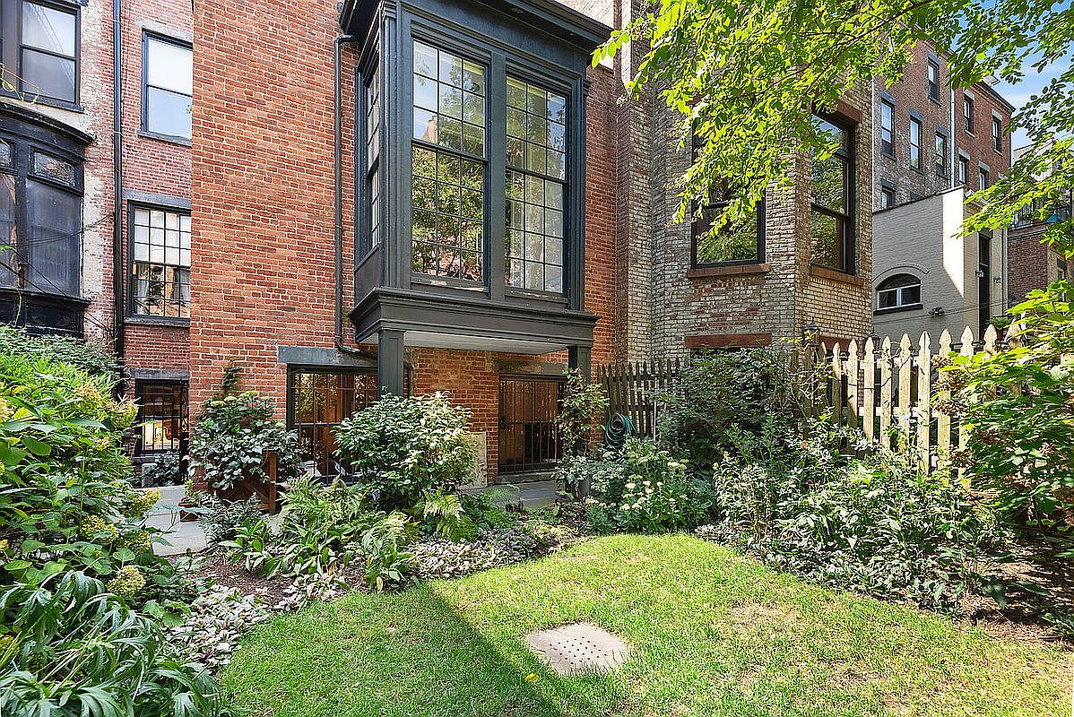 This image captures the rear exterior of a classic brick townhouse, featuring a prominent dark-framed bay window that adds architectural character. A lush, well-maintained garden with a small lawn and dense greenery sits at the base of the building, creating a private and serene outdoor space. The perspective is from the ground level, looking up toward the multi-story structure, emphasizing the blend of historic masonry and natural landscaping.