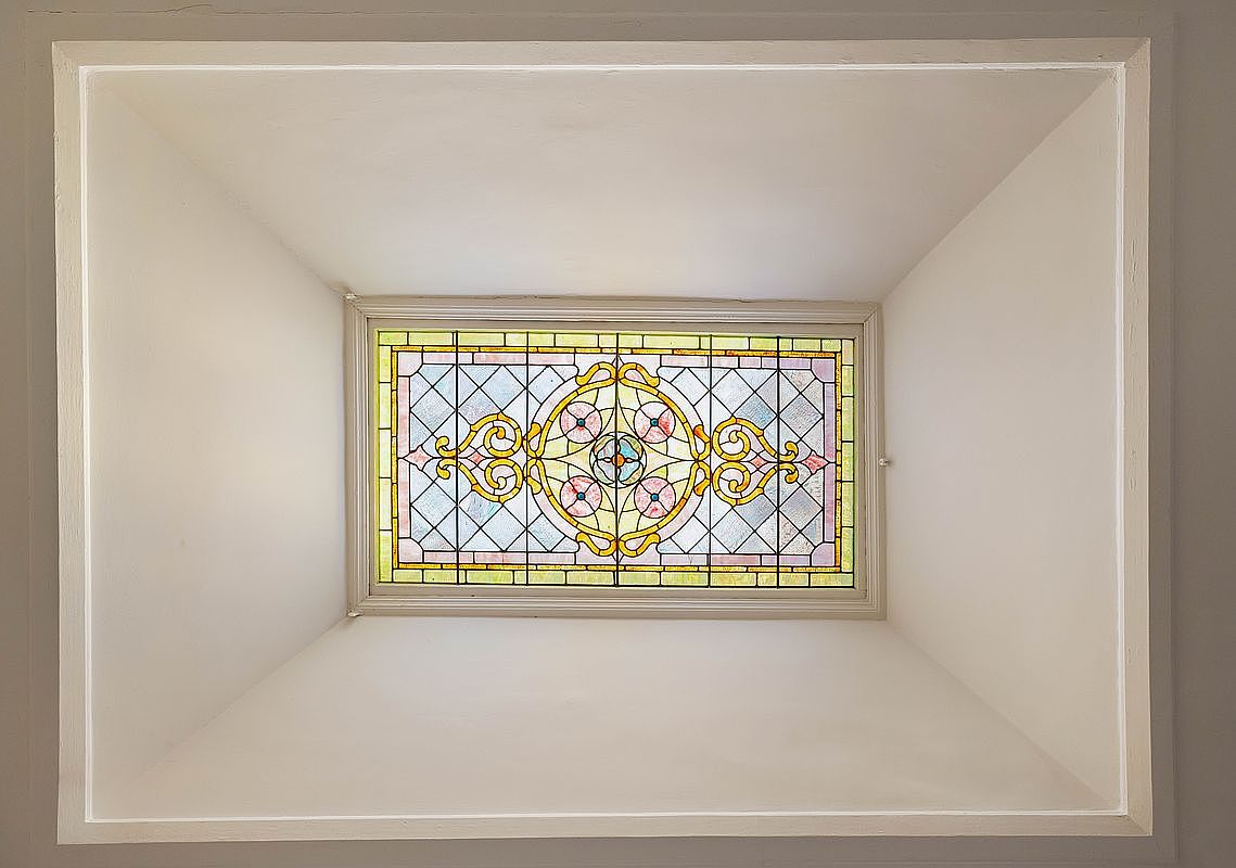 This image captures a stunning stained-glass skylight set within a recessed, white-painted ceiling cove. The intricate, geometric, and floral patterns of the glass cast a soft, diffused light into the space, highlighting the craftsmanship of the architectural detail. The perspective is a direct, upward-facing shot, emphasizing the symmetry and elegant design of this unique ceiling feature.