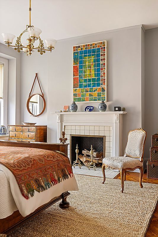 This elegant primary bedroom features a classic fireplace as a focal point, adorned with a vibrant, grid-patterned abstract painting and decorative blue and white vases. A plush, patterned bedspread adds warmth to the room, while a sophisticated upholstered chair and a round hanging mirror enhance the refined, traditional aesthetic. The space is grounded by a textured jute rug and illuminated by a vintage-style brass chandelier, creating a cozy and inviting atmosphere.