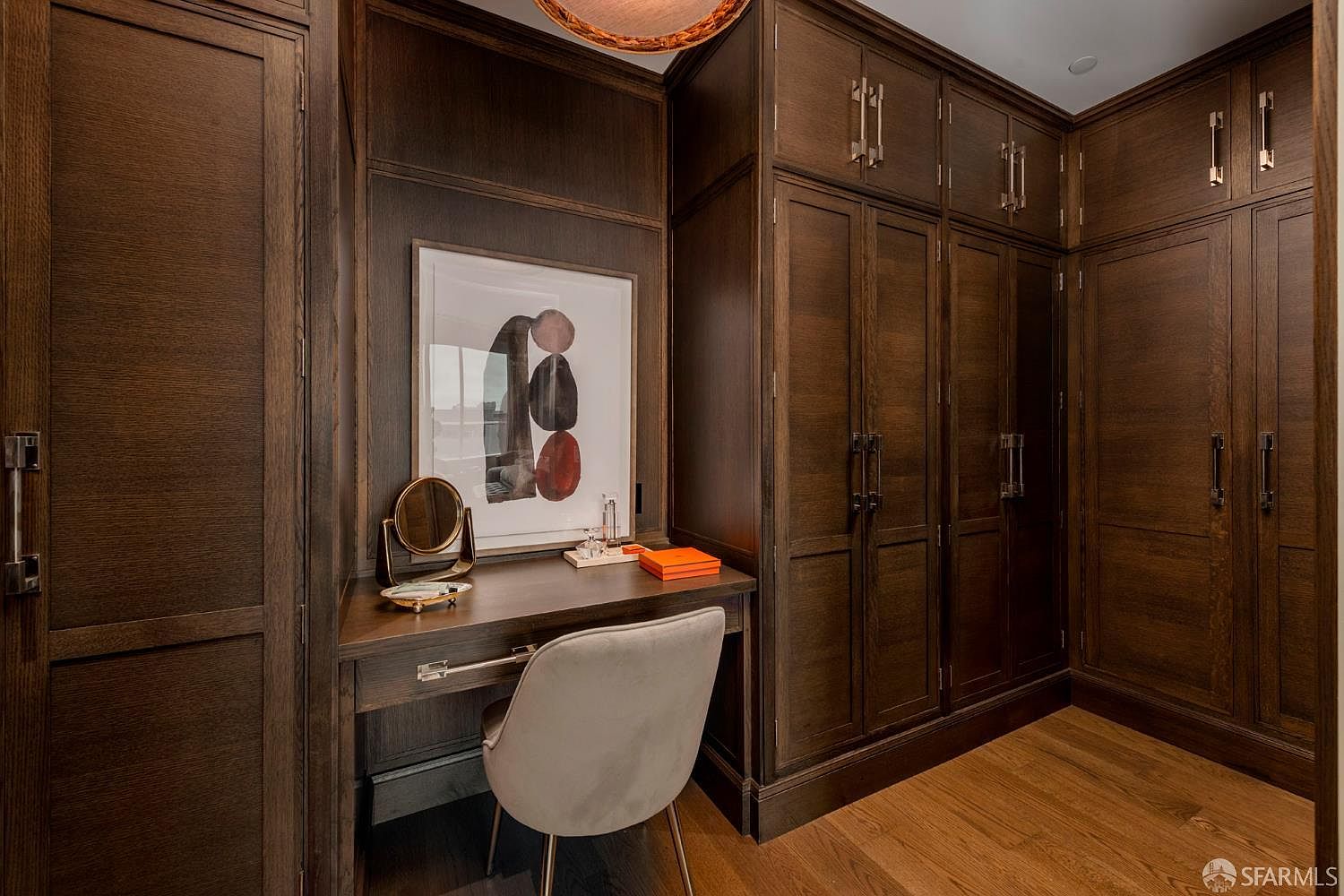 This sophisticated walk-in closet features rich, dark-stained wood cabinetry that provides ample storage space and a built-in vanity area. A modern, neutral-toned chair sits at the desk, which is accented by a contemporary abstract art piece and a small vanity mirror. The warm wood flooring and elegant lighting create a luxurious, high-end atmosphere perfect for a primary suite.