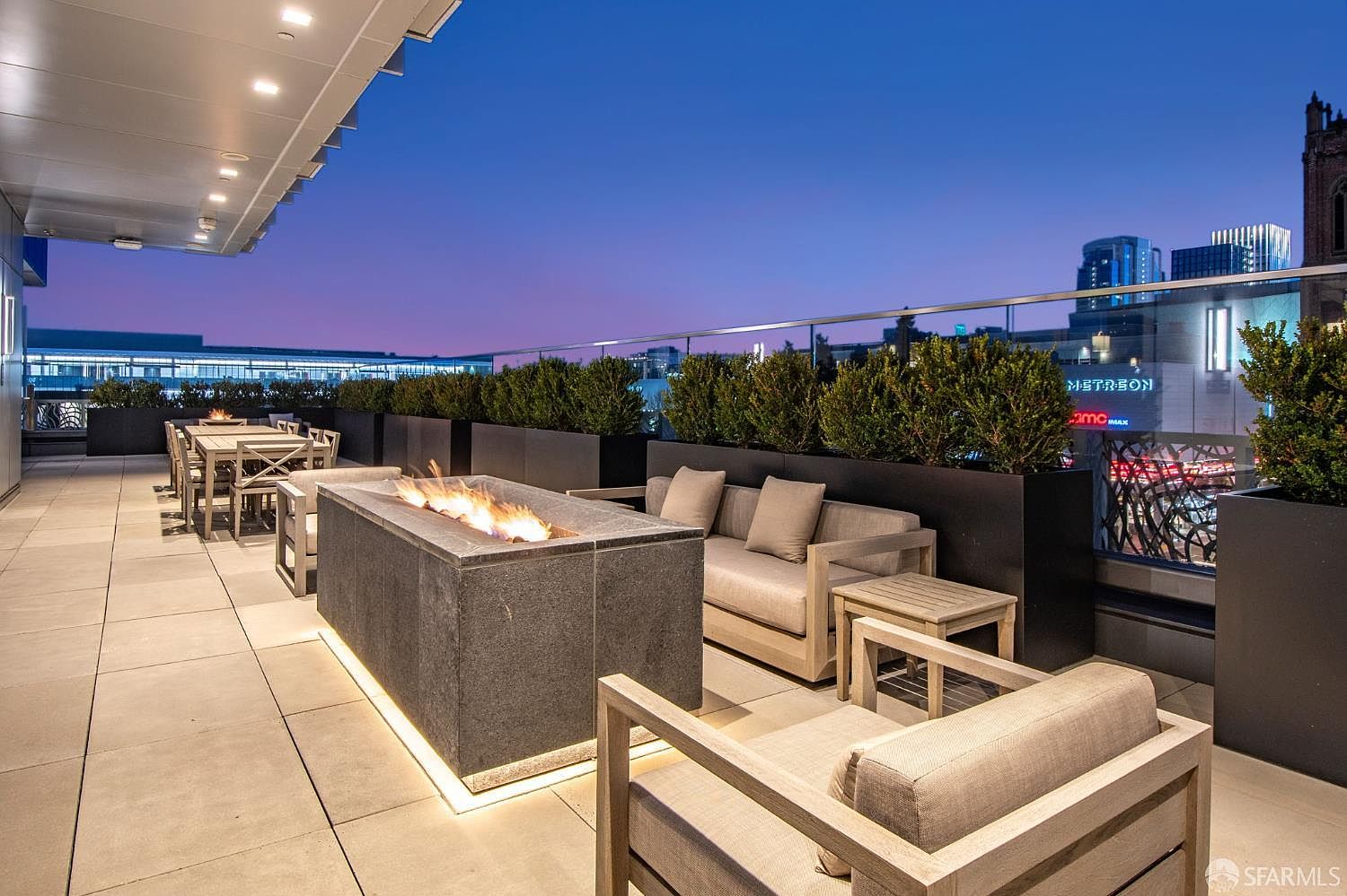 This sophisticated urban rooftop terrace offers a luxurious outdoor living space complete with a modern stone fire pit and comfortable lounge seating. The area is framed by sleek planters filled with greenery and a glass railing that provides an unobstructed view of the city skyline at dusk. The warm glow from the fire pit and under-cabinet lighting creates an inviting, high-end atmosphere perfect for evening entertaining.