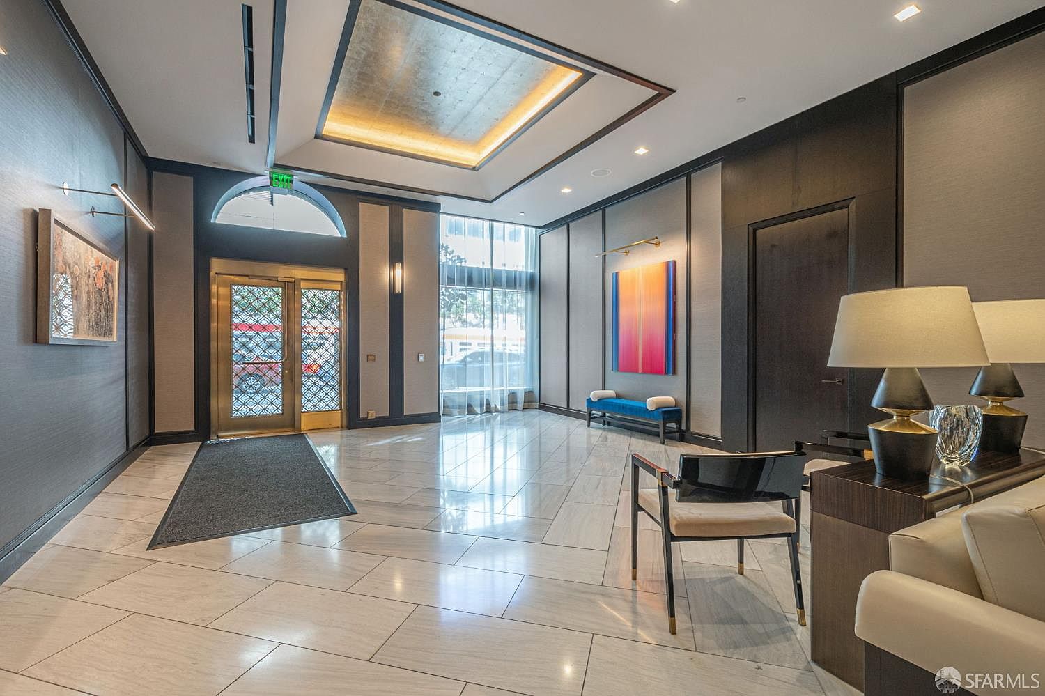 This elegant lobby features polished marble flooring, a dramatic coffered ceiling with recessed lighting, and sophisticated dark wood paneling. The space is accented by a vibrant abstract painting, a sleek blue bench, and a modern chair, creating a refined and welcoming atmosphere for residents and guests. The perspective is a wide-angle shot from the entrance area, emphasizing the grand scale and high-end finishes of the building's common space.
