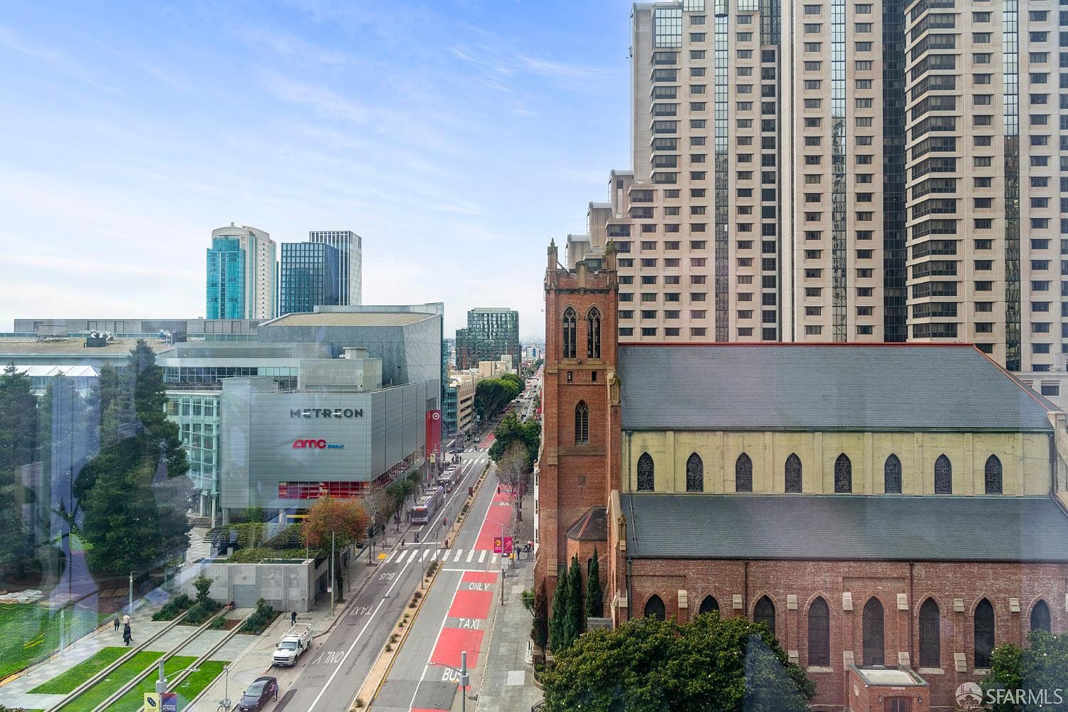 This high-angle aerial view captures a vibrant urban cityscape in San Francisco, featuring a prominent historic brick church adjacent to the modern Metreon shopping and entertainment complex. The perspective looks down onto a busy street with dedicated bus and taxi lanes, framed by towering skyscrapers and clear blue skies. The contrast between the traditional architecture of the church and the contemporary glass structures creates a dynamic and compelling metropolitan scene.