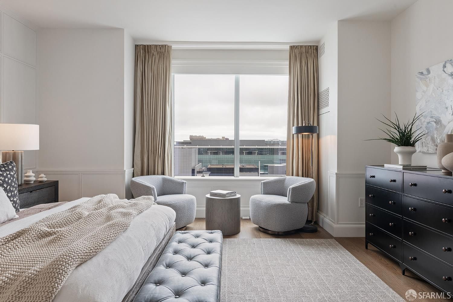 This elegant primary bedroom features a sophisticated neutral color palette, highlighted by a plush tufted bench at the foot of the bed and two cozy swivel chairs positioned by a large window. The room is anchored by a sleek black dresser and a textured area rug, creating a refined and tranquil atmosphere. The large window provides ample natural light and a clear view of the urban cityscape outside.