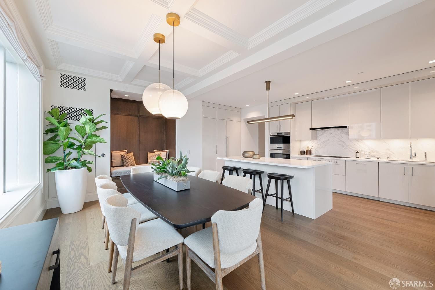 This open-concept space seamlessly blends a modern kitchen with a sophisticated dining area, featuring sleek white cabinetry, a marble-topped island, and a dark wood dining table. The room is illuminated by elegant globe pendant lights and recessed ceiling details, creating a bright and airy atmosphere. A large potted plant adds a touch of greenery, while the warm wood flooring ties the contemporary design together.