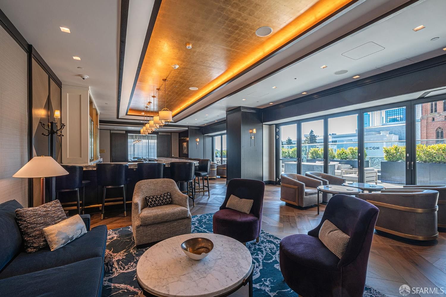 This sophisticated lounge area features a high-end bar with gold-leaf ceiling accents and modern pendant lighting, creating an upscale atmosphere. The space is furnished with plush velvet armchairs, a marble-topped coffee table, and a dark blue patterned rug, all set against a backdrop of floor-to-ceiling glass doors that open onto a private terrace. The overall aesthetic is luxurious and contemporary, perfect for social gatherings or relaxing in a high-rise residential setting.