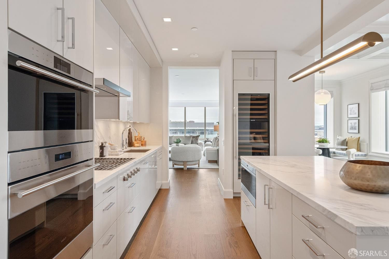 This sleek, modern kitchen features high-gloss white cabinetry, premium stainless steel Wolf appliances, and a spacious marble-topped island. The perspective looks down the galley-style layout toward a bright, open living area, emphasizing the seamless flow and high-end finishes. A minimalist linear pendant light hangs above the island, creating a sophisticated and airy atmosphere.