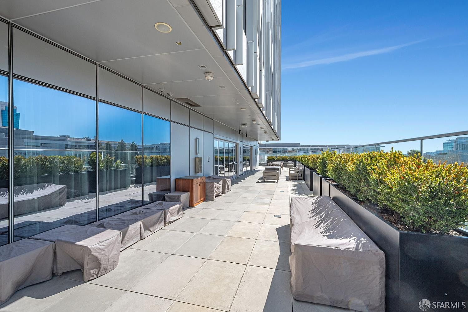 This modern, high-rise outdoor terrace features sleek, minimalist design elements including large-format stone pavers and a long, integrated planter box filled with lush greenery. The space is furnished with several covered outdoor seating arrangements, offering a clean and sophisticated area for relaxation against a backdrop of clear blue skies and urban architecture. The perspective is a wide-angle shot looking down the length of the balcony, emphasizing the expansive and well-maintained nature of this premium amenity space.