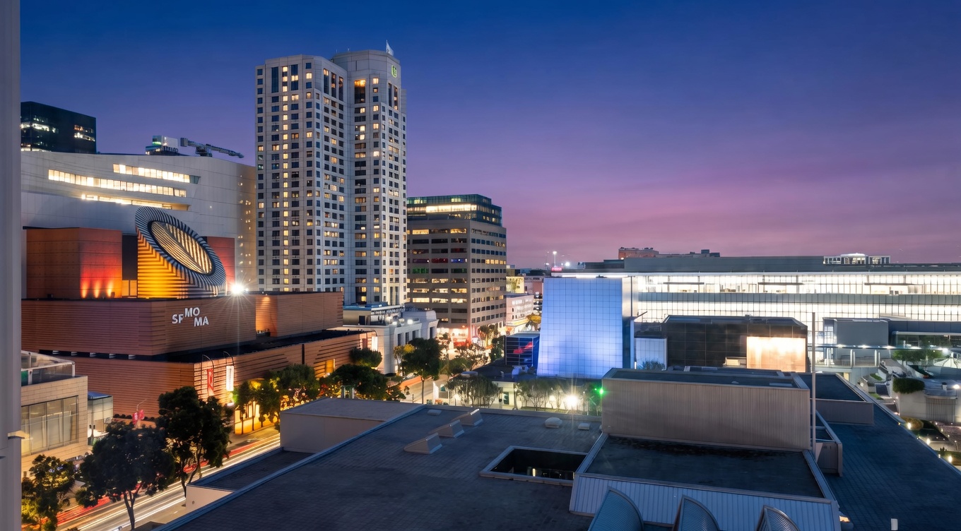 This stunning high-angle exterior view captures the San Francisco Museum of Modern Art (SFMOMA) and the surrounding urban skyline during twilight. The perspective highlights the architectural design of the building, including its iconic circular oculus, against a vibrant purple and blue evening sky. The shot features well-integrated city amenities, street lighting, and a dense cityscape that conveys a sophisticated, metropolitan living environment.