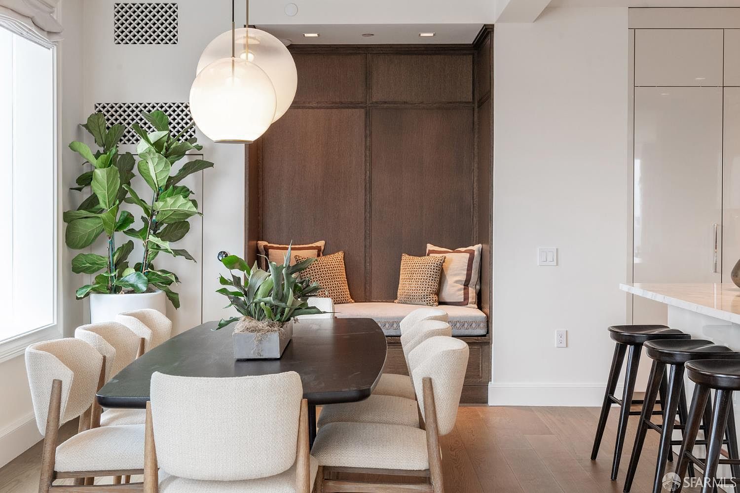 This sophisticated dining area features a dark, organic-shaped table surrounded by modern, cream-colored upholstered chairs. A built-in wooden banquette with plush cushions provides cozy seating against a dark wood-paneled wall, while a large fiddle leaf fig plant adds a touch of natural greenery. The space is illuminated by elegant, spherical pendant lights, creating a warm and inviting atmosphere perfect for refined entertaining.