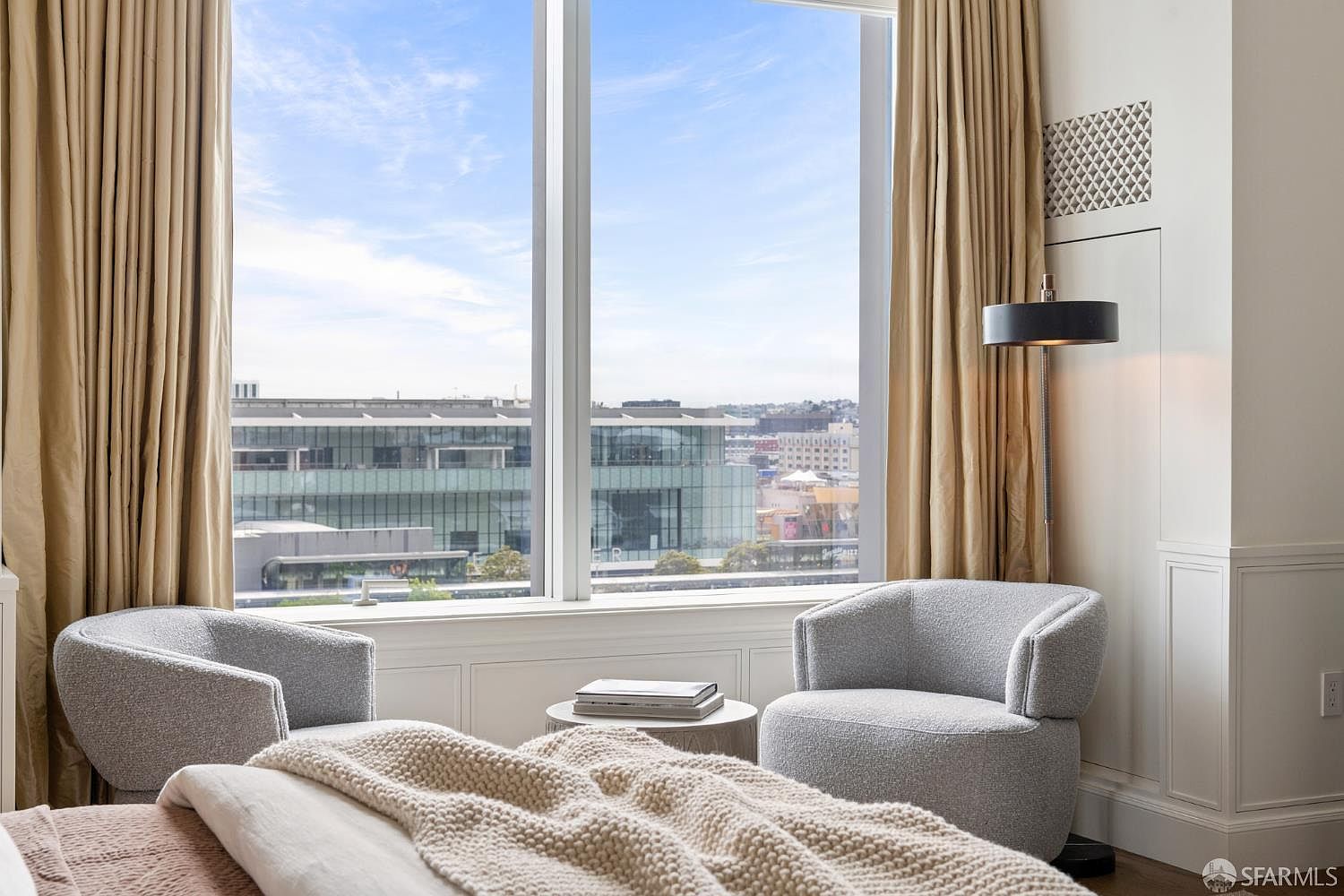 This elegant bedroom features a cozy seating area with two plush, light gray armchairs positioned by a large floor-to-ceiling window that offers a sweeping urban view. A textured, cream-colored knit throw blanket rests on the bed in the foreground, while a sleek floor lamp and neutral-toned curtains add a sophisticated, tranquil atmosphere to the space. The room exudes a refined, modern luxury aesthetic, perfect for a high-end residential listing.
