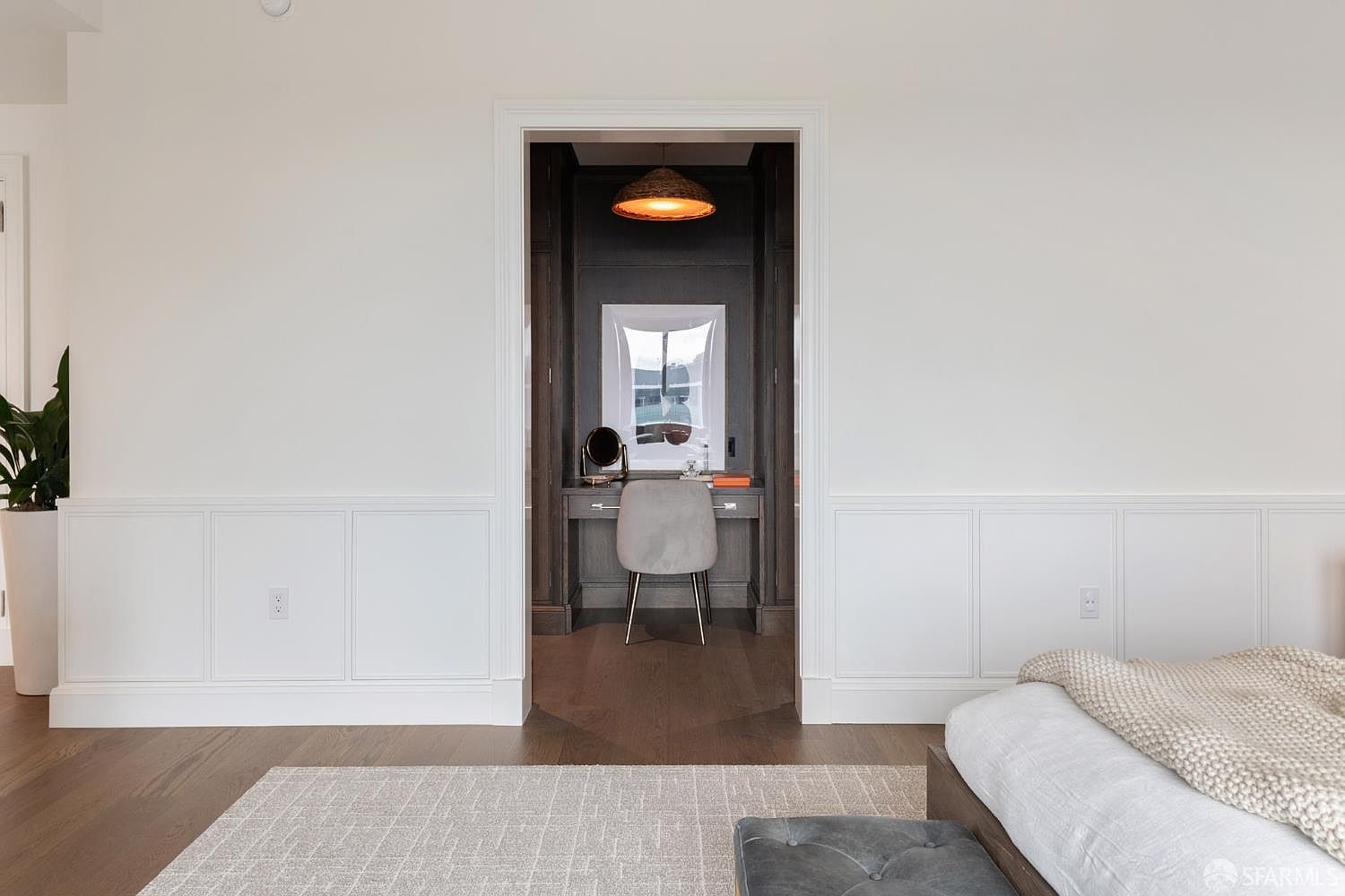 This image captures a view from a primary bedroom looking into a sophisticated, dark-toned walk-in closet or dressing area. The foreground features a neutral-toned bedroom with white wainscoting and a plush rug, while the framed doorway reveals a dedicated vanity space with a modern chair, warm pendant lighting, and built-in cabinetry. The contrast between the bright, airy bedroom and the moody, intimate dressing nook creates a luxurious and well-designed atmosphere.