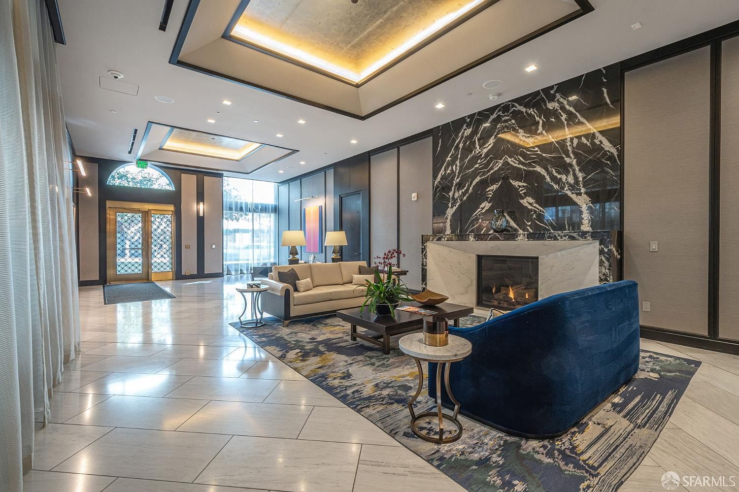 This elegant lobby or lounge area features a sophisticated blend of modern luxury and classic design, highlighted by a striking black marble fireplace surround and a coffered ceiling with recessed lighting. A plush blue velvet sofa and a neutral-toned sofa are arranged on a large, patterned area rug, creating a welcoming and upscale seating area. The space is characterized by high ceilings, polished marble floors, and a grand entryway, conveying a sense of prestige and comfort suitable for a high-end residential building.