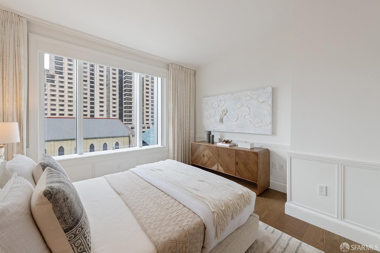 This bright and airy primary bedroom features a neutral color palette, a plush bed with textured pillows, and a warm wooden sideboard topped with minimalist decor. A large window provides an expansive urban view, while elegant wainscoting and soft drapery add a touch of sophisticated charm to the space. The room exudes a serene and upscale atmosphere, perfect for a modern city residence.