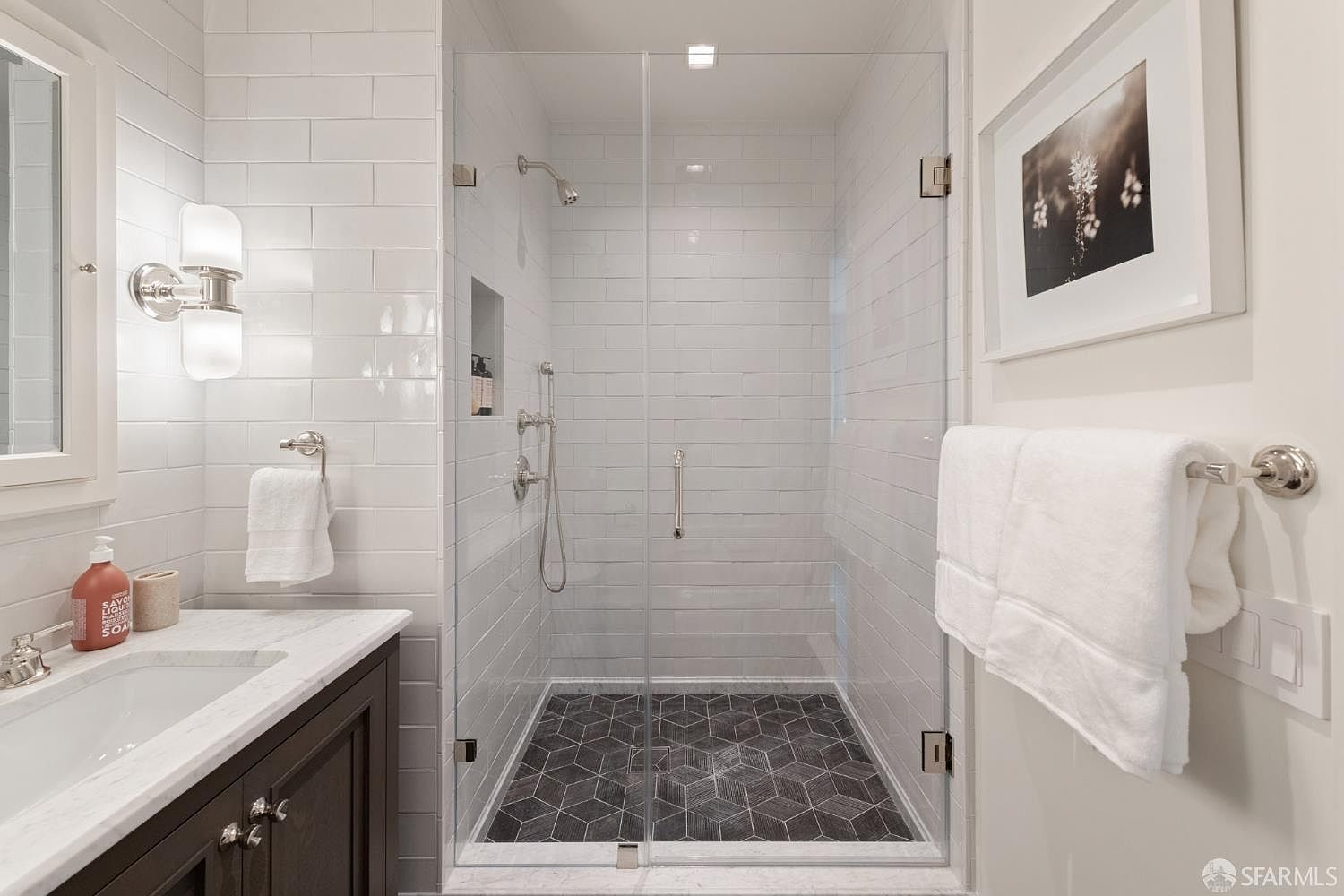 This elegant bathroom features a walk-in shower with a glass enclosure, white subway tile walls, and a striking dark geometric tile floor. A dark wood vanity with a marble countertop and a modern wall-mounted light fixture adds a sophisticated touch to the space. The room is bright and clean, showcasing a contemporary design with high-quality finishes.