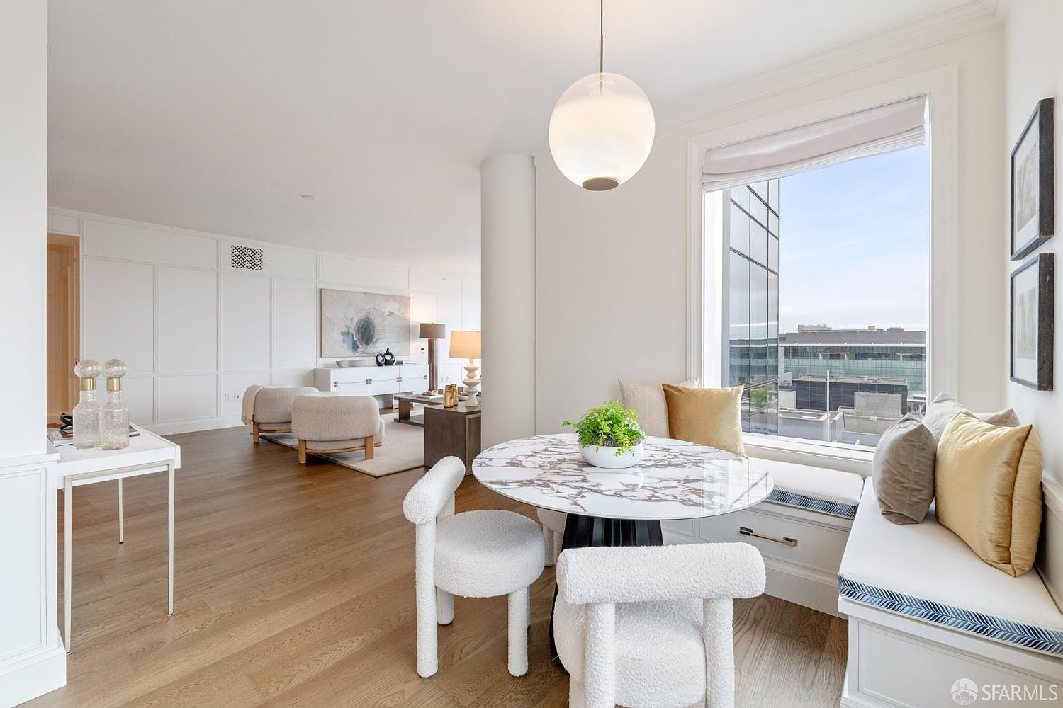 This bright and airy dining area features a modern marble-topped table paired with textured, cream-colored chairs and a built-in window bench with plush gold and grey pillows. The space is illuminated by a contemporary globe pendant light and offers a seamless transition into an open-concept living room with light hardwood floors and elegant wall paneling. The overall aesthetic is sophisticated and minimalist, emphasizing natural light and high-end finishes.