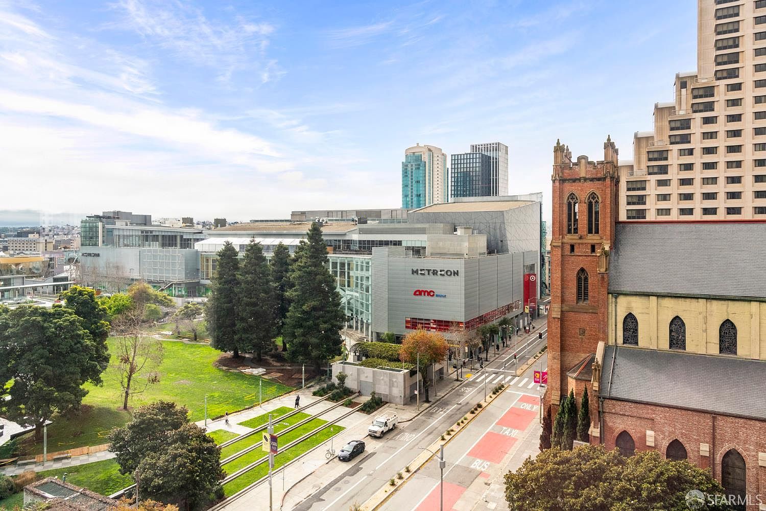 This high-angle aerial view captures a vibrant urban scene in San Francisco, featuring the prominent Metreon shopping center and AMC theater complex adjacent to a historic brick church. Below, a lush green park with walking paths provides a peaceful contrast to the bustling city street, which is marked with bus and taxi lanes. The perspective offers a comprehensive look at the neighborhood's blend of modern commercial architecture and traditional landmarks under a bright, expansive sky.