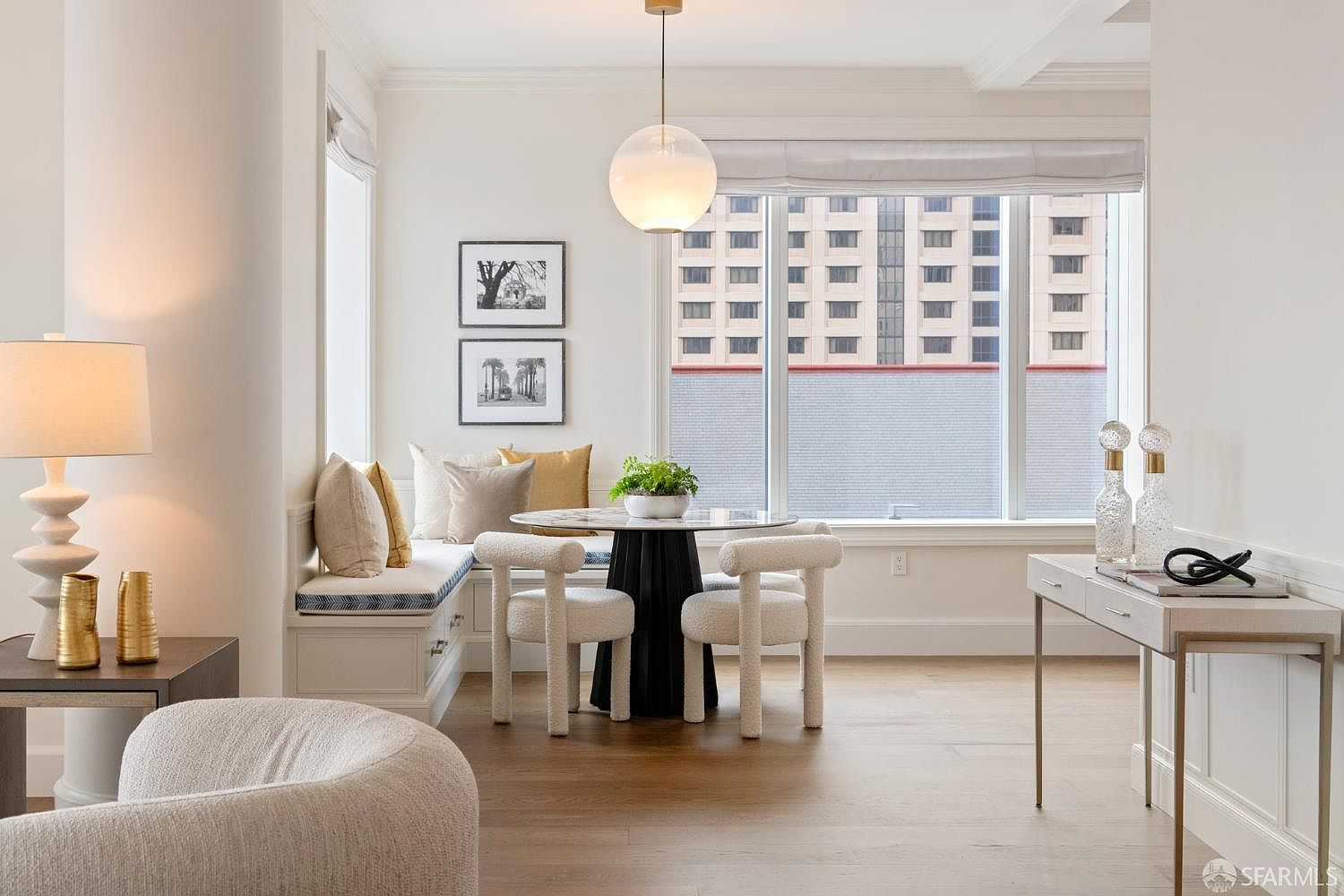 This elegant dining area features a cozy built-in banquette with plush cushions, paired with a modern round marble-top table and two textured, cream-colored chairs. A sleek, spherical pendant light hangs above the table, while large windows provide ample natural light and a view of the city skyline. The space is accented by framed artwork and a minimalist console table, creating a sophisticated and inviting atmosphere.