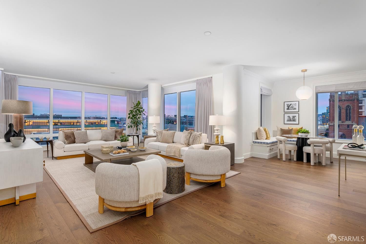 This expansive, light-filled living room features a sophisticated open-concept layout with floor-to-ceiling windows offering panoramic city views. The space is anchored by a neutral color palette, plush cream-colored seating, and warm hardwood flooring, creating an inviting and luxurious atmosphere. A dining nook with built-in bench seating and a modern pendant light completes the elegant, high-end aesthetic of this urban residence.