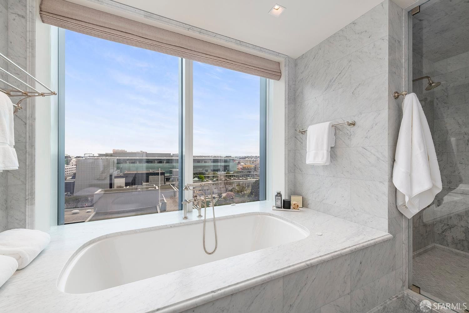 This luxurious primary bathroom features a deep soaking tub set against a large window that offers an expansive urban view. The space is finished in elegant, light-toned marble tile throughout, creating a bright and sophisticated atmosphere. A glass-enclosed shower and high-end fixtures complete the modern, spa-like aesthetic.