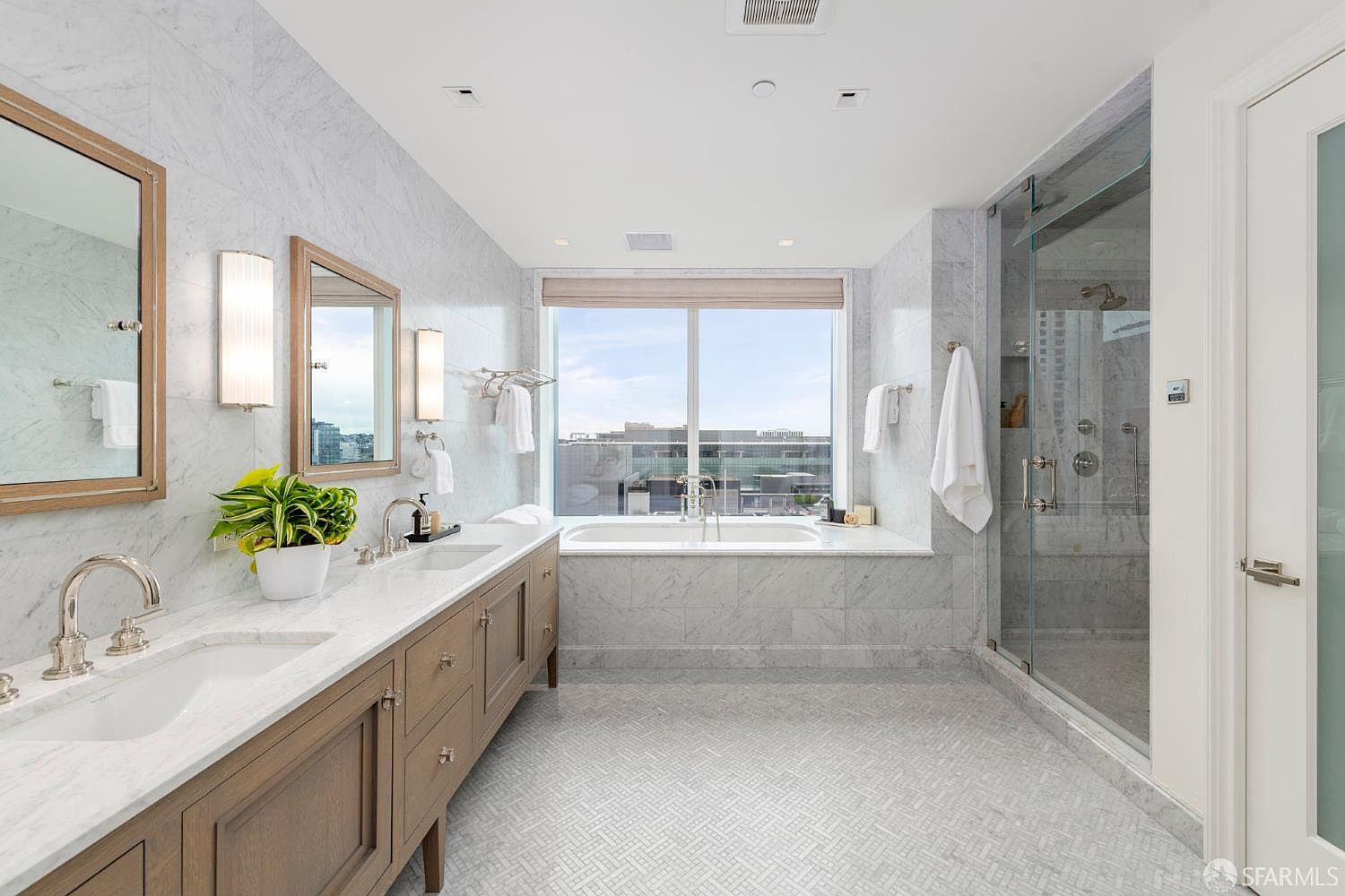 This luxurious primary bathroom features a double vanity with wood cabinetry and marble countertops, complemented by elegant wall sconces and framed mirrors. A deep soaking tub is positioned beneath a large window that offers expansive city views, while a spacious walk-in glass shower with marble tiling completes the high-end aesthetic. The room is bathed in natural light, creating a serene and sophisticated spa-like atmosphere.