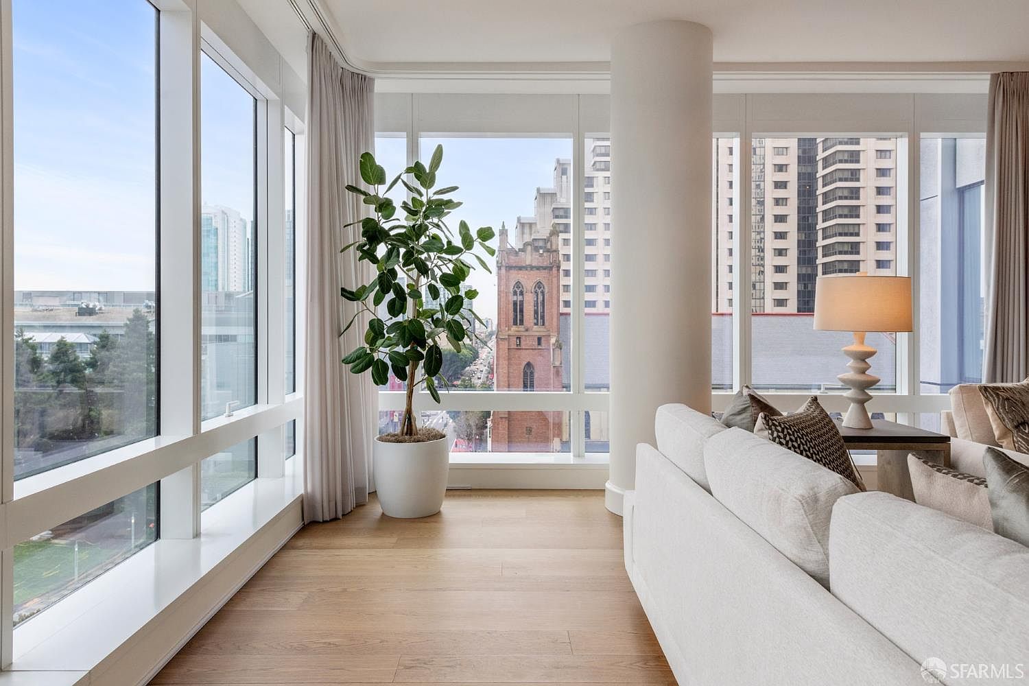 This bright and airy living room features floor-to-ceiling windows that offer a stunning urban view, including a historic church and modern high-rises. The space is anchored by a plush, light-colored sofa and a large potted plant, creating a serene and sophisticated atmosphere. The minimalist design and natural light emphasize the luxury and openness of the high-rise residence.