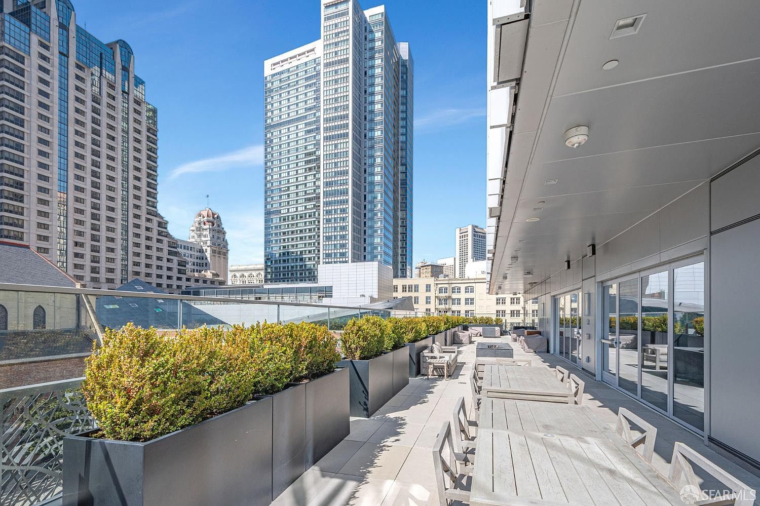 This expansive, modern rooftop terrace offers a sophisticated outdoor living space with a long dining table and comfortable seating areas. The deck is lined with sleek, dark metal planters filled with lush greenery, providing a vibrant contrast against the backdrop of towering city skyscrapers. The perspective captures the urban luxury of the space, emphasizing the seamless blend of contemporary design and metropolitan views.