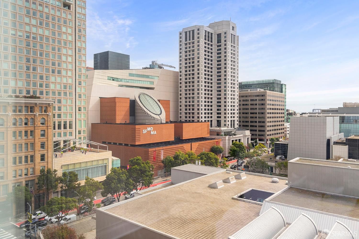 This high-angle aerial view captures a vibrant urban cityscape in San Francisco, prominently featuring the iconic SFMOMA building with its distinctive brick facade and circular skylight. The surrounding area is densely packed with modern high-rise office buildings, residential towers, and tree-lined streets, showcasing a bustling metropolitan environment. The perspective provides a clear sense of the property's central location within a major cultural and commercial hub.