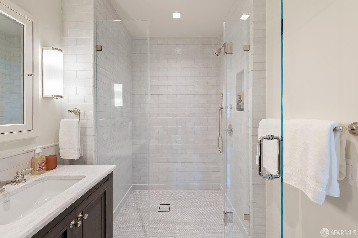 This clean and modern bathroom features a walk-in shower with white subway tile walls and a mosaic tile floor, enclosed by a frameless glass door. A dark wood vanity with a white marble countertop and a sleek faucet sits to the left, complemented by a wall-mounted light fixture and a white towel hanging nearby. The overall aesthetic is bright, minimalist, and well-maintained, offering a crisp and inviting atmosphere.