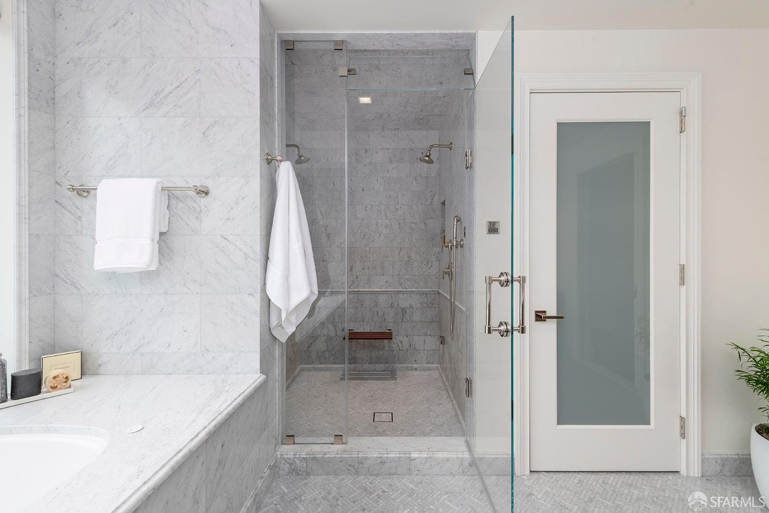 This luxurious primary bathroom features a spacious walk-in shower enclosed by frameless glass, showcasing elegant marble tiling throughout the walls and floor. A built-in wooden shower bench and dual showerheads add a spa-like feel, while a frosted glass door provides privacy and a modern aesthetic. The bright, clean design is complemented by high-end finishes and a marble-topped vanity area, creating a sophisticated and serene atmosphere.