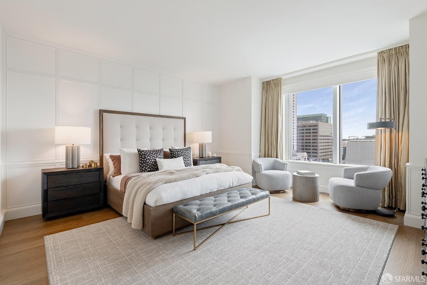 This elegant primary bedroom features a sophisticated neutral color palette, highlighted by a large upholstered headboard set against a classic white paneled wall. The room is furnished with a plush bed, a tufted grey bench, and two modern armchairs positioned by a floor-to-ceiling window that offers an expansive urban view. The space is grounded by a textured area rug and warm wood flooring, creating a serene and luxurious atmosphere.