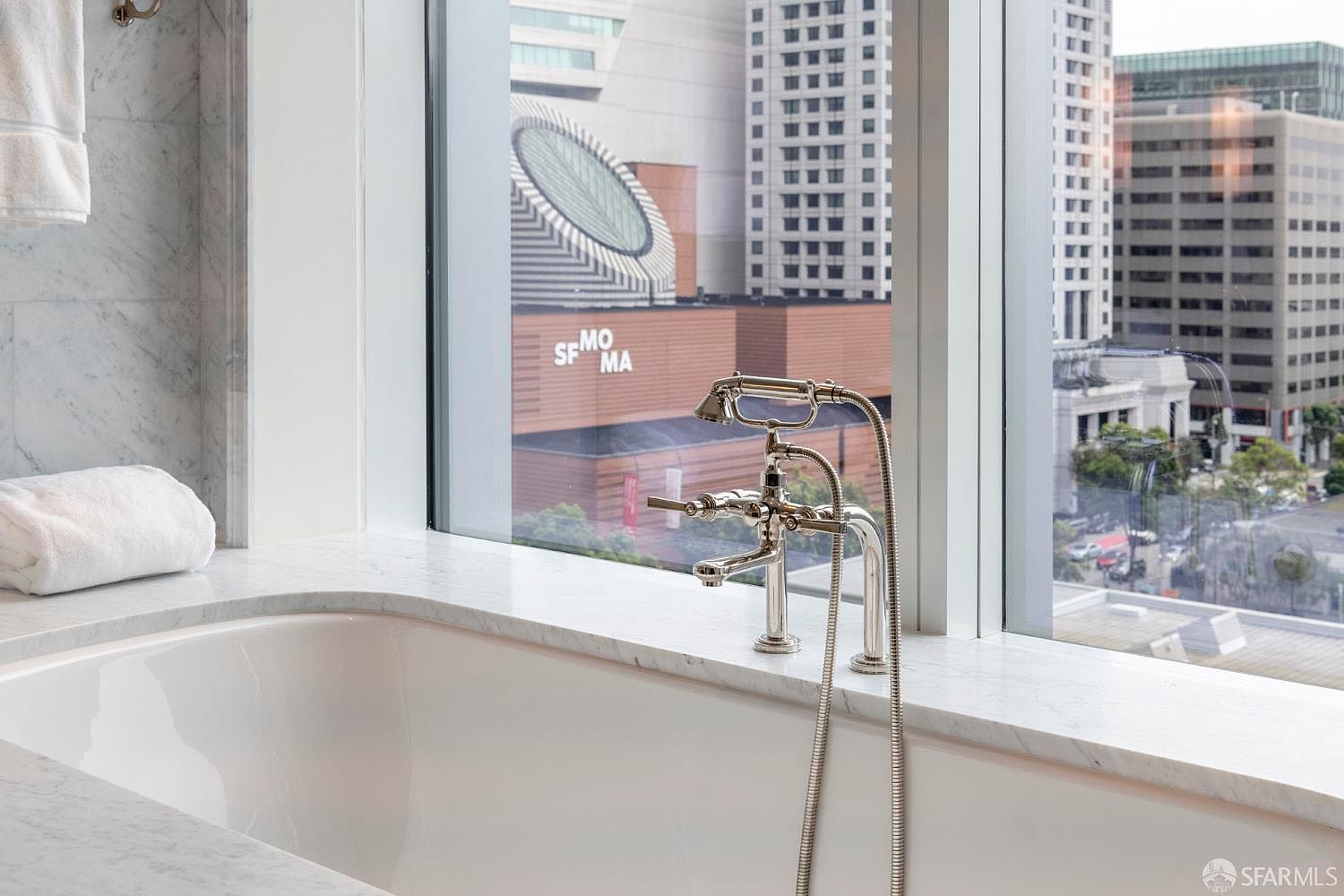 This elegant bathroom features a deep soaking tub set against a large window that offers a striking, direct view of the SFMOMA building and the surrounding city skyline. The space is finished with high-end marble surfaces and a polished chrome faucet fixture, creating a luxurious and serene atmosphere. The perspective highlights the seamless blend of sophisticated interior design and an iconic urban backdrop.