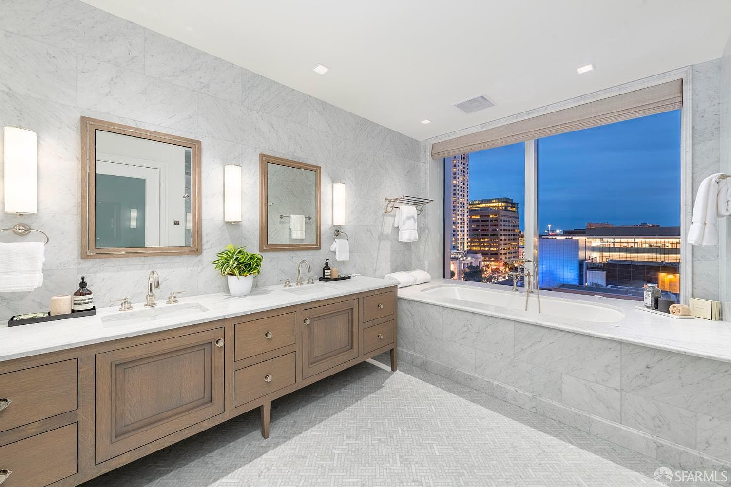 This luxurious primary bathroom features a double vanity with light wood cabinetry and marble countertops, complemented by elegant wall-mounted mirrors and sconces. A deep soaking tub is positioned perfectly beneath a large window that offers a stunning, expansive view of the city skyline at dusk. The room is finished with sophisticated marble wall tiles and a detailed mosaic floor, creating a serene and high-end spa-like atmosphere.