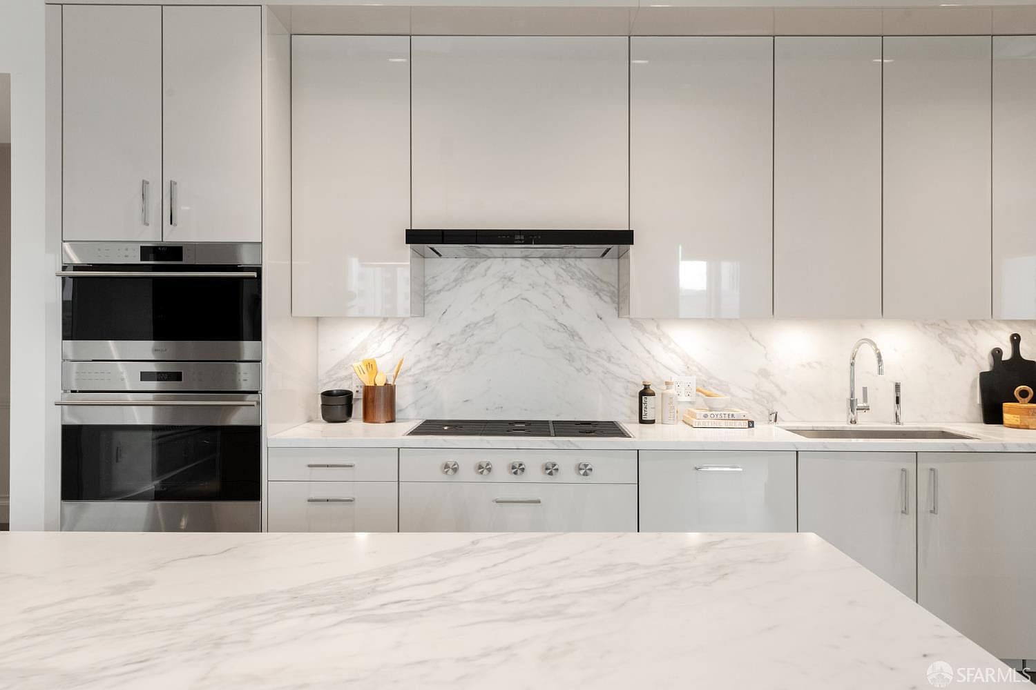 This sleek, modern kitchen features high-gloss white cabinetry paired with a striking marble backsplash and matching countertop. The space is equipped with premium stainless steel appliances, including a built-in wall oven and a gas cooktop, creating a clean and sophisticated aesthetic. The perspective is a straight-on, eye-level shot that highlights the seamless integration of the appliances and the bright, minimalist design.