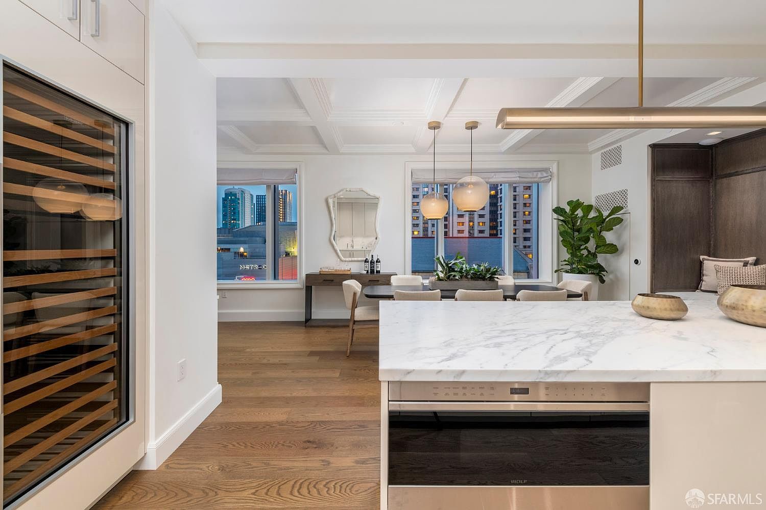 This sophisticated, open-concept kitchen features a large marble-topped island with a built-in Wolf oven, seamlessly transitioning into a bright dining area. The space is characterized by high-end finishes, including a tall wine refrigerator, warm hardwood flooring, and elegant pendant lighting. Large windows provide an expansive urban view, creating a luxurious and airy atmosphere perfect for modern living.