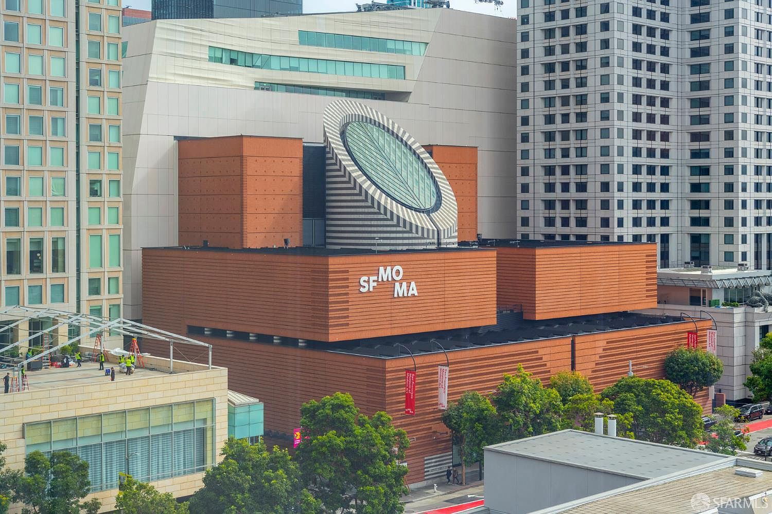 This high-angle aerial shot captures the iconic San Francisco Museum of Modern Art (SFMOMA), showcasing its distinctive brick-clad facade and the prominent, circular, striped skylight structure. The building is nestled within a dense urban environment, surrounded by modern glass skyscrapers and lush green trees lining the street below. The perspective emphasizes the architectural contrast between the museum's bold, geometric design and the surrounding city skyline.