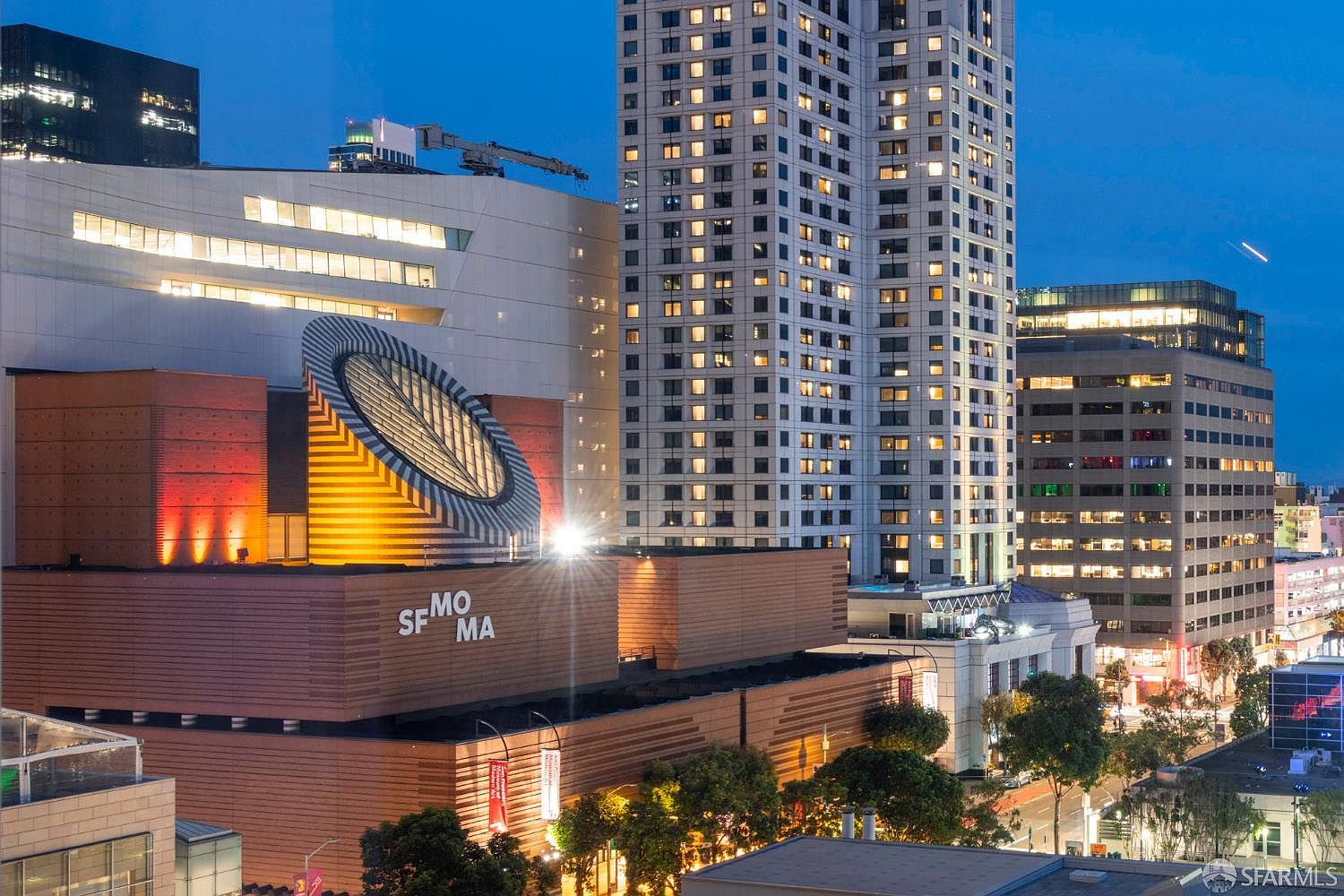This high-angle aerial shot captures the iconic SFMOMA building in San Francisco at twilight, showcasing its distinctive modern architecture, including the prominent circular skylight feature. The composition highlights the museum's relationship with the surrounding urban skyline, featuring tall residential towers and dense city lights. The warm glow from the building's facade and the surrounding skyscrapers creates a sophisticated and vibrant metropolitan atmosphere.