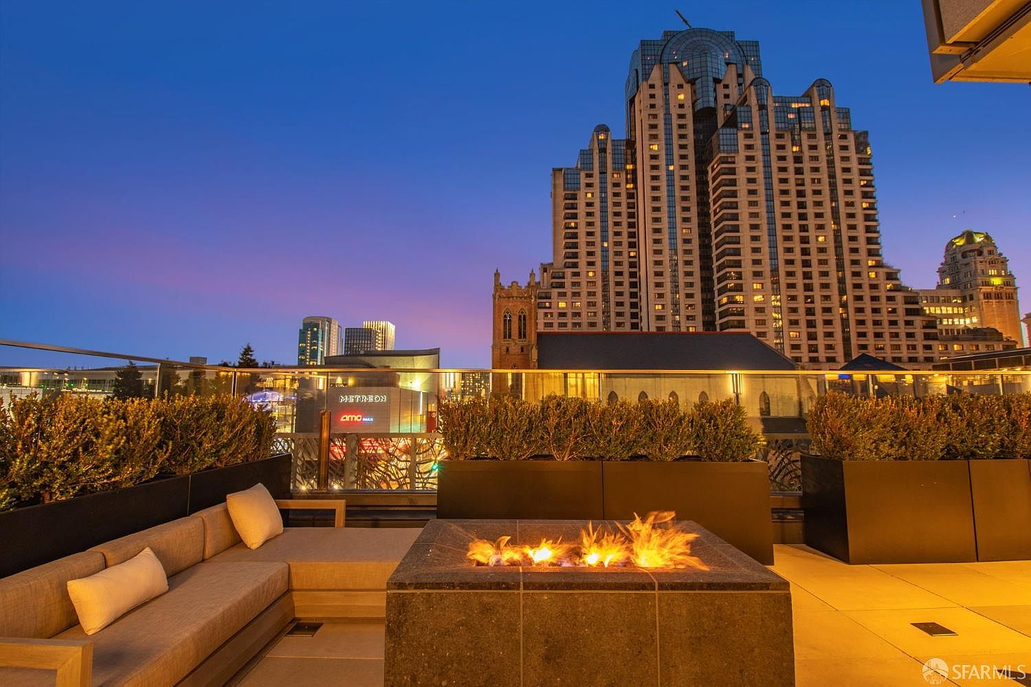 This sophisticated rooftop terrace offers a stunning panoramic view of the city skyline at dusk, featuring a modern, square stone fire pit as the central focal point. A comfortable, contemporary sectional sofa with plush cushions provides an inviting seating area for outdoor entertaining. The space is framed by sleek glass railings and manicured hedges, creating a luxurious and serene urban retreat.
