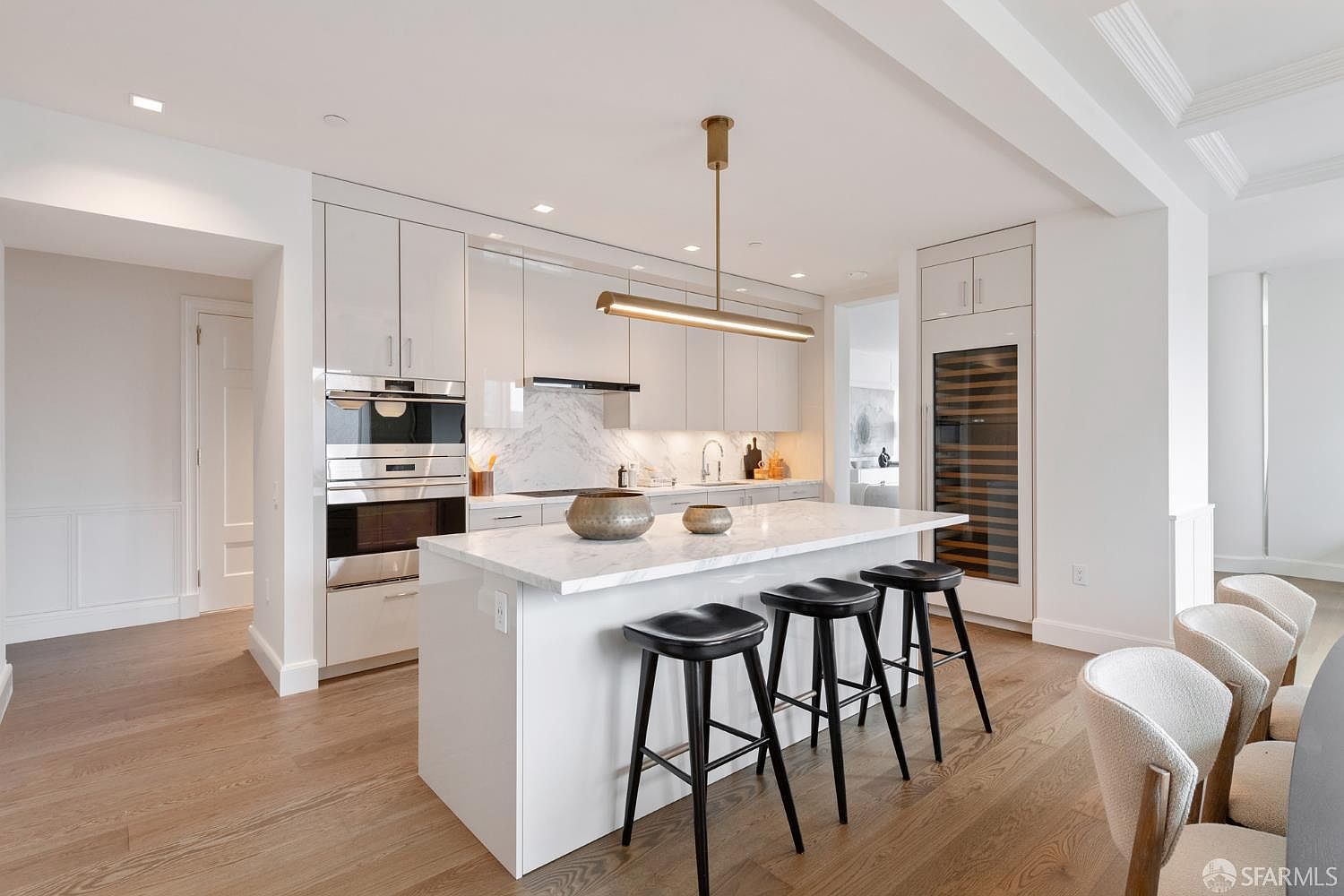 This sleek, modern kitchen features high-gloss white cabinetry, a marble-topped island with three black bar stools, and premium stainless steel appliances. A minimalist linear pendant light hangs above the island, while a built-in wine refrigerator adds a touch of luxury to the bright, open-concept space. The light wood flooring and clean lines create an airy, sophisticated atmosphere perfect for contemporary living.