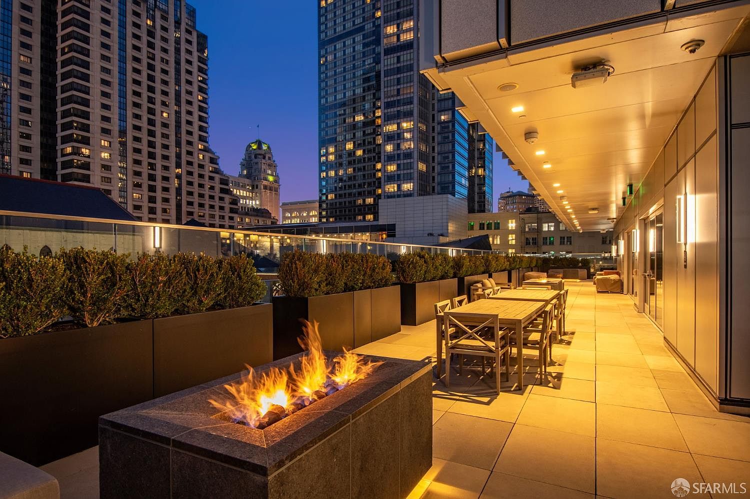 This sophisticated urban terrace offers a stunning evening view of the city skyline, featuring a modern fire pit as a central focal point. The space is furnished with a long dining table and chairs, bordered by sleek planters filled with lush greenery, creating an inviting atmosphere for outdoor entertaining. The warm, ambient lighting under the building's overhang enhances the luxurious and tranquil mood of this high-rise balcony.