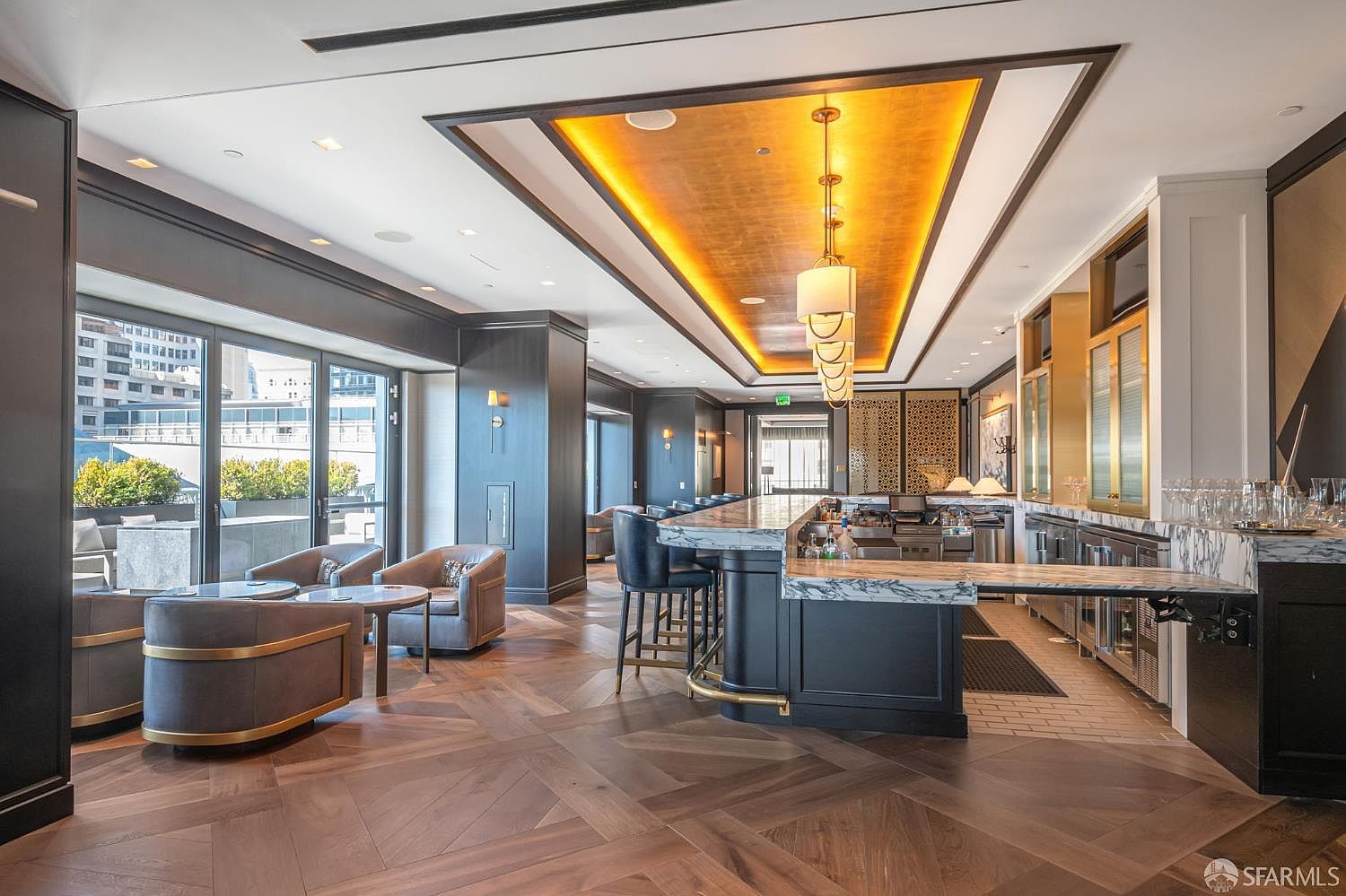 This sophisticated lounge area features a high-end bar with a marble countertop, dark cabinetry, and elegant gold-leaf ceiling accents. The space is furnished with plush, modern armchairs and bar stools, all set against a backdrop of rich herringbone wood flooring and floor-to-ceiling glass doors that open to an outdoor terrace. The overall aesthetic is one of refined luxury, perfect for social gatherings in a premium residential building.
