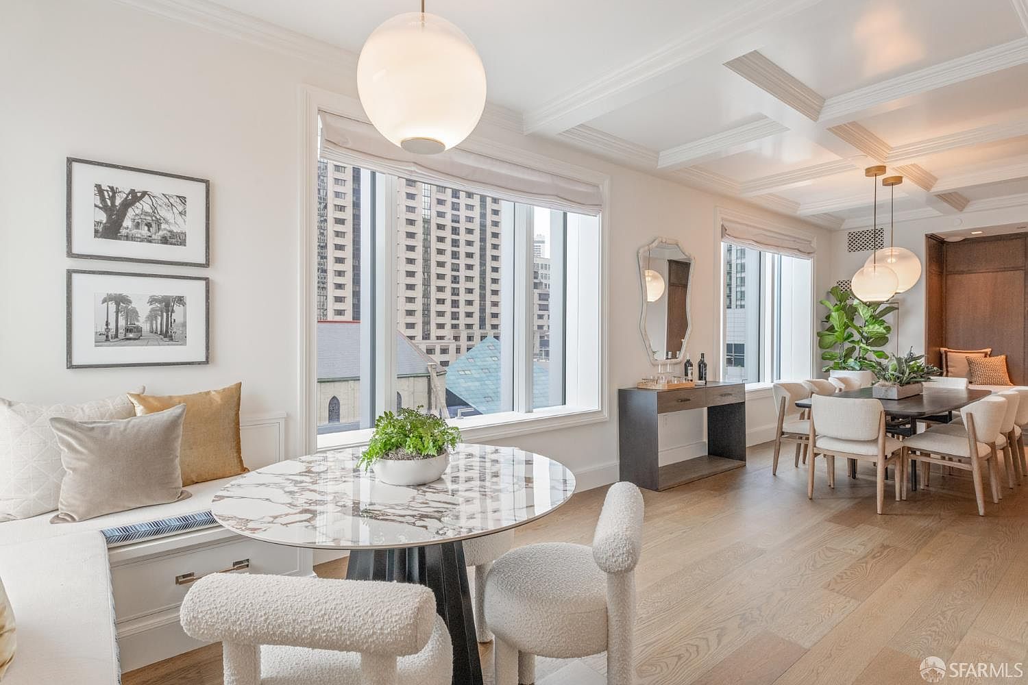 This elegant dining area features a bright, open-concept layout with a marble-topped breakfast nook and a formal dining table set for guests. The space is characterized by sophisticated modern furniture, light hardwood flooring, and a coffered ceiling, all bathed in natural light from large windows overlooking a city skyline. The neutral color palette and curated decor create a refined, airy atmosphere perfect for both casual meals and formal entertaining.