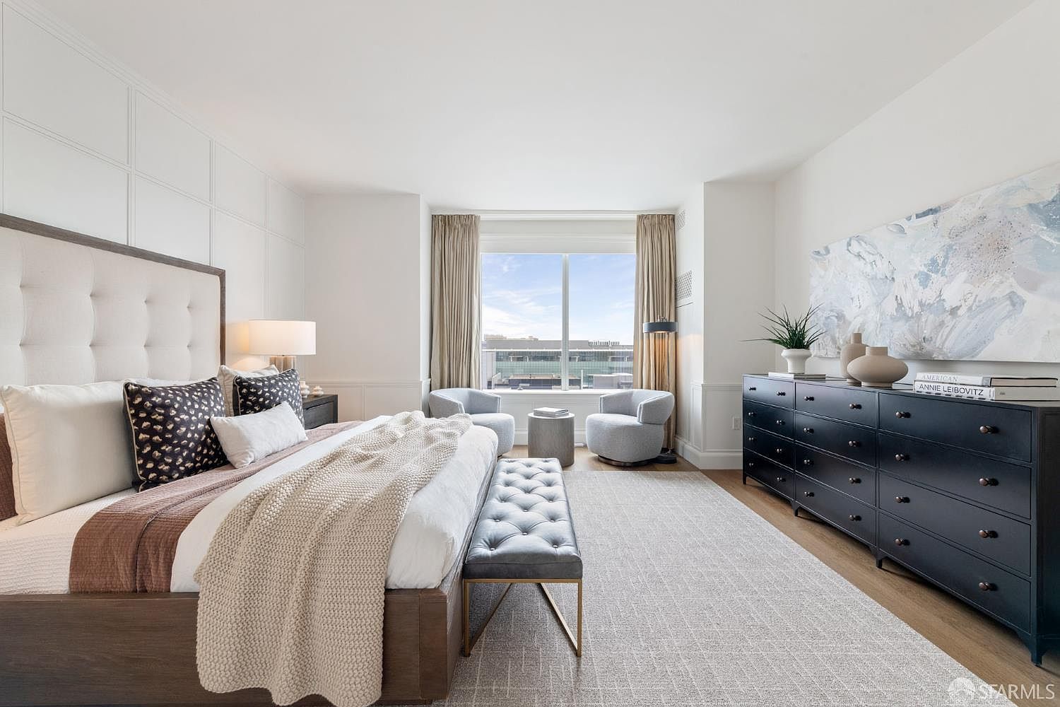This elegant primary bedroom features a large, tufted upholstered headboard and a sophisticated dark wood dresser, creating a refined and tranquil atmosphere. A cozy seating area with two plush armchairs is positioned by the window, which offers a bright, expansive view of the urban surroundings. The room is anchored by a textured area rug and accented with neutral tones, providing a luxurious and comfortable living space.